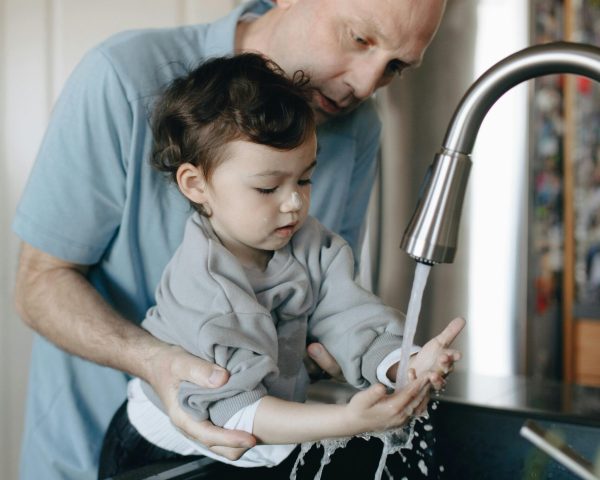 A father tenderly helps his child wash hands in the home kitchen sink.